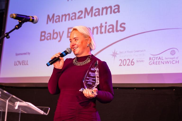 A woman in a burgundy dress holds a glass trophy and speaks into a microphone on stage. Behind her is a screen that reads: Mama2Mama Baby Essentials 