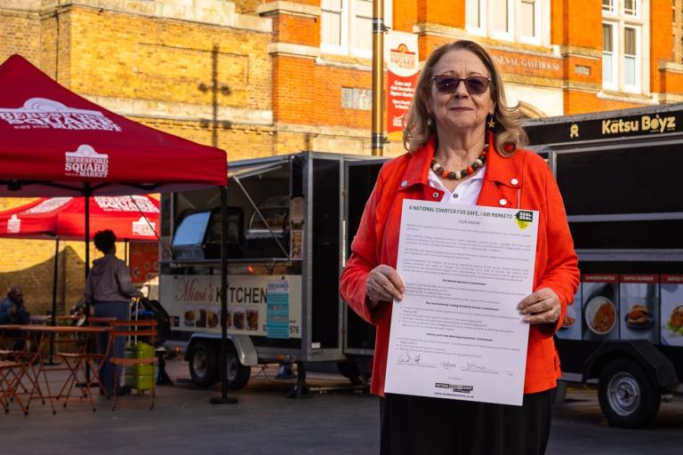 A woman in an orange jacket holds a charter in a market. Behind her are food trucks. In front of one of the trucks is a red gazebo.