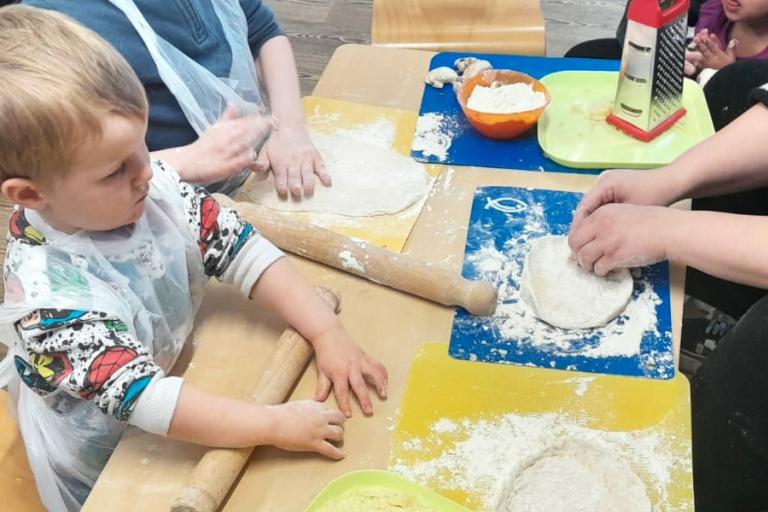 A little boy in an apron helping to roll out some dough with the help of some adults. 