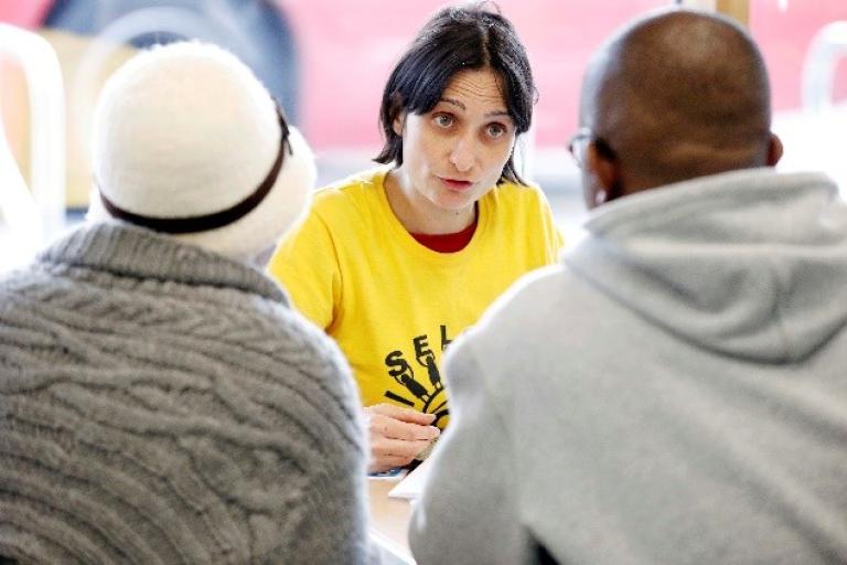 A woman in a yellow t-shirt is sat down talking to two people with their backs to the camera