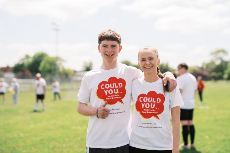 Two people wearing could you foster with the royal borough of greenwich t-shirts - smiling to camera while stood on a football pitch