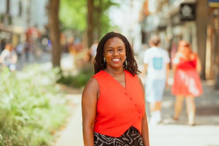 Kaia is wearing a bright orange top standing on a tree-lined street in Woolwich town centre.