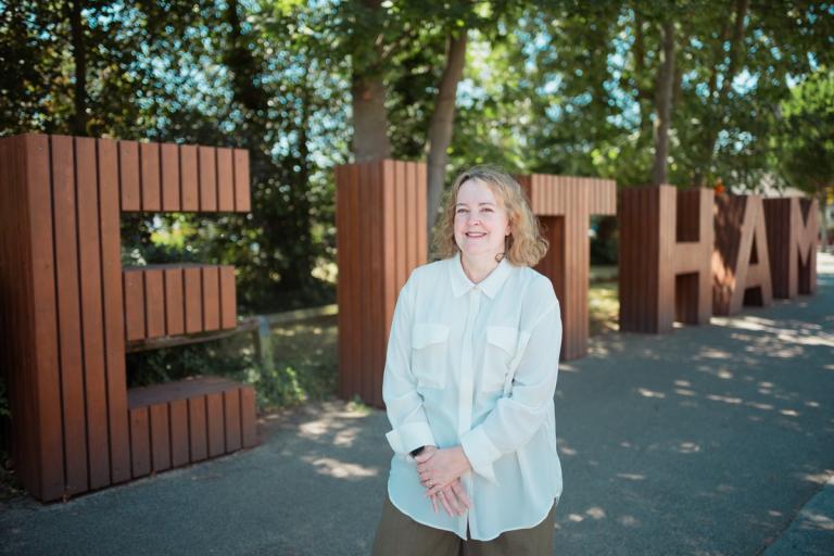 Wendy standing outdoors in front of large wooden ELTHAM sign with trees and a shaded pathway in the background.