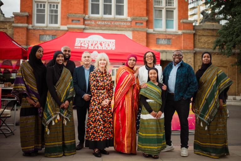 Group photo of Cllr Adel Khaireh, Lord Len Duvall, Deputy Mayor of London Justine Simons OBE, Cllr Asli Mohammed and Cllr Anthony Okereke with Somali dancers in traditional dress in Beresford Square Market standing in front of a red market stall and the Royal Arsenal Gatehouse.