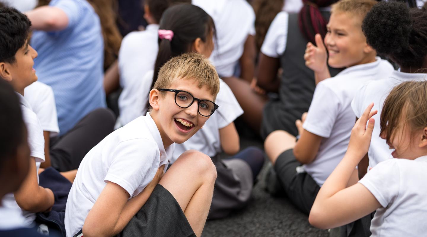 Children sitting down in a playground, one child in glasses is looking directly at the camera smiling.