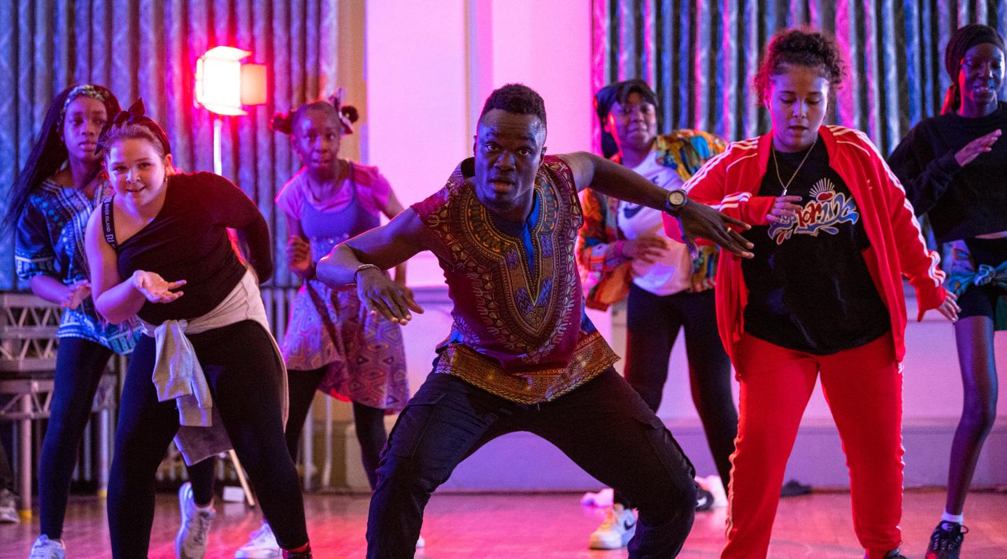 A lively dance class with diverse participants in colorful outfits, energetically performing in a well-lit room with purple and red lighting accents.