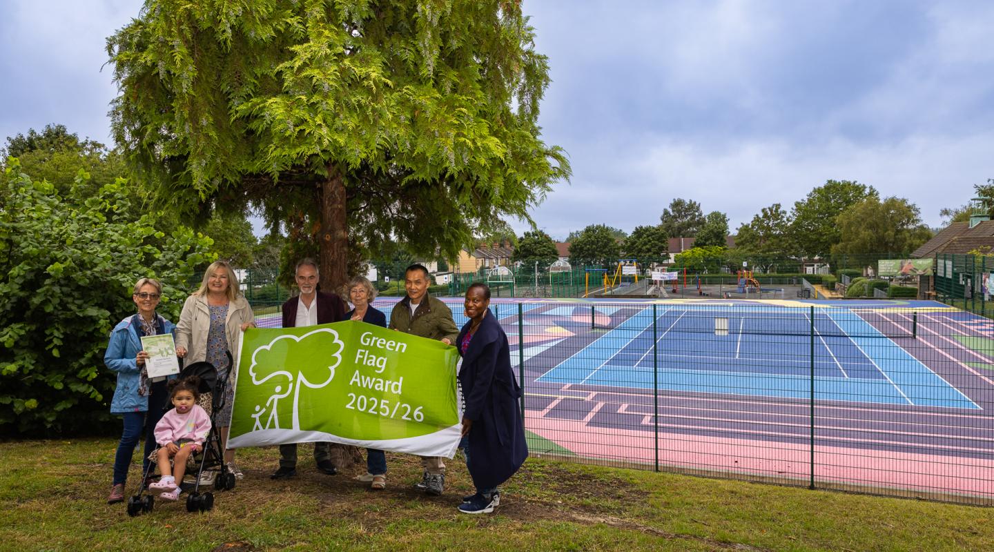 A group of people standing on grass holding a flag that reads 'Green Flag 25/26' A big tree is behind them and a tennis court beyond that.