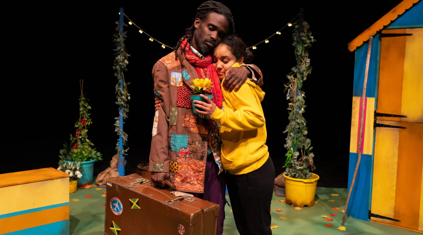Two people hugging. One is wearing a brown coat and carrying a leather suitcase with Jamaican flags on. The other is wearing a yellow hoodie.