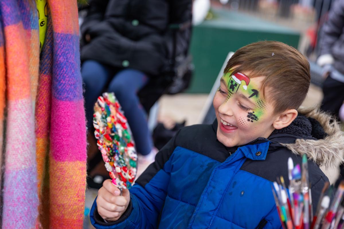 A child with green face paint on smiling into a handheld mirror