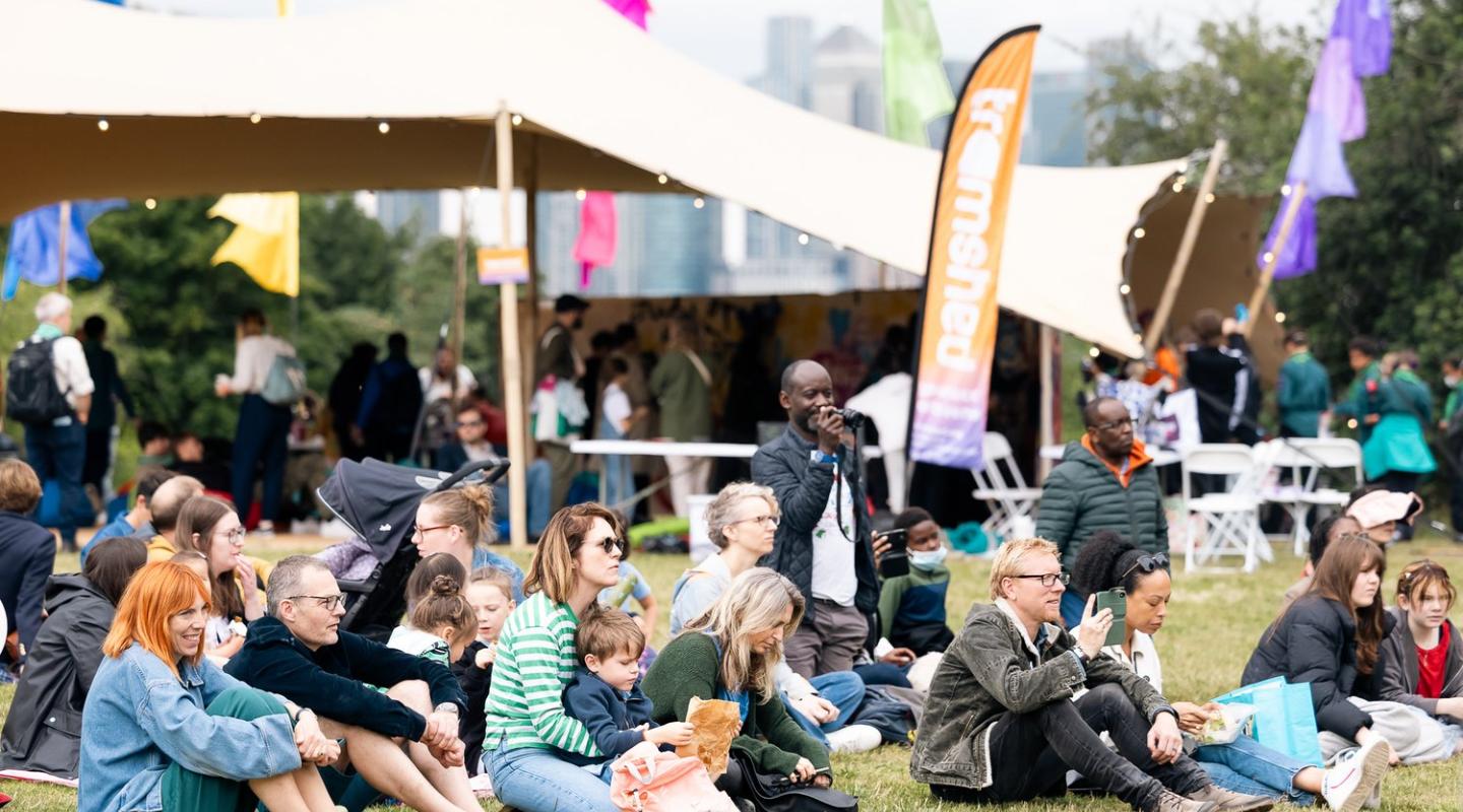 A group of people sitting down on grass at an outdoors event. Behind them is an open air tent and a flag that says 'tramshed'